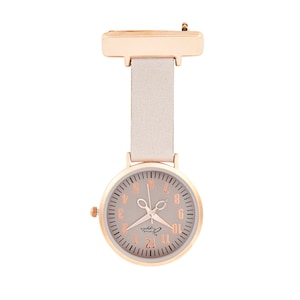 Puede incluir: Un reloj de bolsillo de enfermera en oro rosa con esfera y correa gris claro. La esfera del reloj presenta números y manecillas en oro rosa, con un diseño de tijeras. El texto "Annie Apple" está impreso en la esfera.