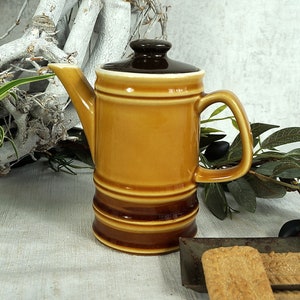 Op de afbeelding: Een bruine keramische kan met een bruin deksel en een brede handgreep. De kan heeft een bruin en beige gestreept ontwerp. De kan staat op een lichtbruin oppervlak.