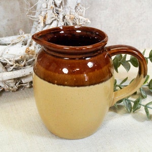 Könnte beinhalten: Ein brauner und beiger Keramik-Krug mit rundem Körper und Henkel. Der Krug hat eine braune Glasur auf der oberen Hälfte und eine beige Glasur auf der unteren Hälfte.