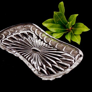 Op de afbeelding: Een heldere, rechthoekige kristallen schaal met een zonnestraalontwerp in het midden. De schaal heeft een geschulpte rand en wordt vergezeld door een takje groene bladeren tegen een zwarte achtergrond. Ideaal voor het serveren van kleine items.