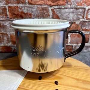 May include: A silver metal coffee maker with a white ceramic filter and a black handle. The coffee maker has the text "Melitta 103" printed on the side.