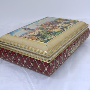 Può includere: Una scatola vintage in latta rossa e oro con un design trapuntato e una scena di una città europea sul coperchio.