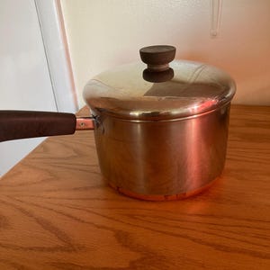 May include: A copper-colored saucepan with a matching lid and a dark brown handle. The lid has a dark brown knob. The saucepan is sitting on a wooden surface.