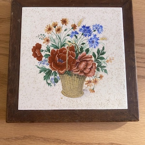 May include: A vintage ceramic tile with a wooden frame features a colorful floral arrangement in a woven basket. The flowers include red, orange, yellow, blue, and green hues.