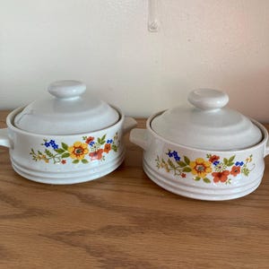 May include: Two white ceramic lidded bowls with floral designs. Each bowl has a handle on either side and a small knob on the lid. The floral pattern includes yellow, orange, blue, and green flowers and leaves.