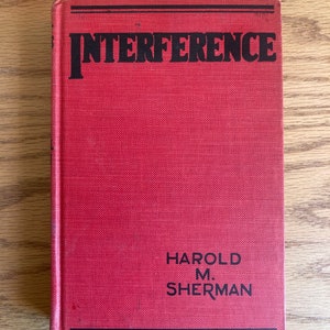 May include: A red hardcover book with the title "INTERFERENCE" in black bold letters and the author's name "HAROLD M. SHERMAN" in black letters at the bottom.