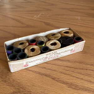 May include: A vintage box of Star Darning and Mending thread. The box contains multiple spools of thread in various colors, including gold, black, red, and purple. The box has text that reads "Star Darning and Mending".
