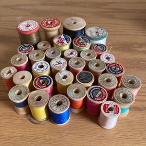 May include: A collection of vintage wooden spools of thread in various colors, including red, blue, green, yellow, and pink. The spools are labeled with different brands, including Coats and Clarks.