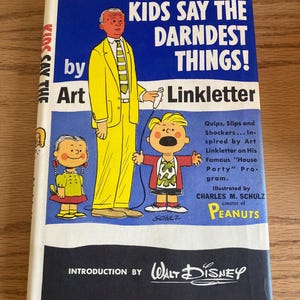 May include: Book cover with a blue background and the title "Kids Say the Darndest Things!" by Art Linkletter. The cover features illustrations of Art Linkletter, Charlie Brown, and Lucy from the Peanuts comic strip. The book is illustrated by Charles M. Schulz, creator of Peanuts.