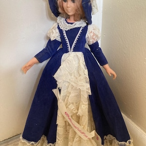 May include: A vintage doll wearing a blue velvet dress with white lace trim and a matching bonnet. The doll has blonde hair and is holding a ribbon with text. The dress has long sleeves and a full skirt.