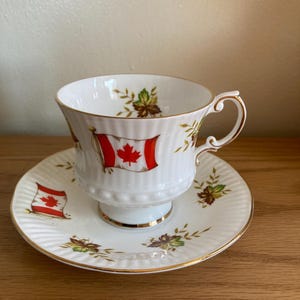 May include: A white teacup and saucer with a gold rim. The teacup has a red and white Canadian flag design and green leaves. The saucer has a similar design with a smaller flag.