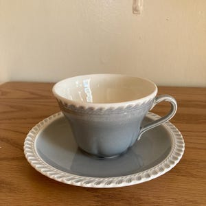 May include: A ceramic teacup and matching saucer set featuring a soft blue-grey glaze. The cup has a curved handle and a cream-colored interior. Both pieces are finished with a decorative, raised rope-like texture along the outer rim edges.