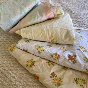 May include: A collection of folded vintage bed sheets in various floral patterns. The sheets feature pastel colors, including yellow, pink, green, and white. Some sheets have rose and floral designs, while others have striped patterns.