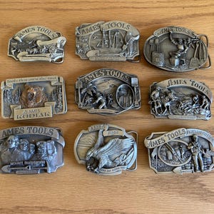 May include: Nine silver belt buckles with various designs featuring the Ames Tools logo. The buckles feature images of a bear, an eagle, a mountain, and a soldier. The text on the buckles includes "Tools that own the turf", "Mount Rushmore", "Eagle", "Fights for Freedom", and "Ames Tools".