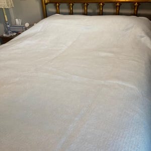 May include: A solid white bedspread covers a mattress, featuring a smooth, uniform texture across the surface. The bedspread is positioned against a metallic gold-toned headboard with vertical spindles and horizontal bars at the top and bottom.