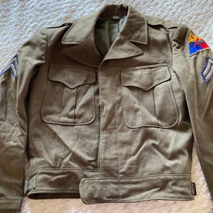 May include: A vintage military jacket in olive green with a patch on the right shoulder and a chevron on the left sleeve. The jacket has two large pockets on the front and a belt at the waist.