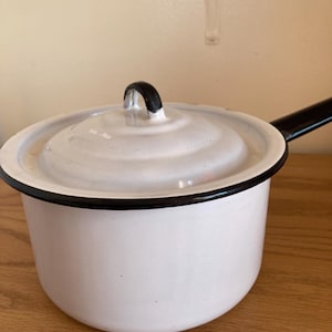 May include: White enamel saucepan with a black handle and rim. The pot has a lid with a black handle. The saucepan is sitting on a wooden surface. Kitchenware.