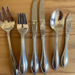 May include: Silverware set including a serving fork, dinner fork, two knives, a spoon, and a butter knife. The handles have a teardrop shape with a beaded edge. The utensils are arranged on a wooden surface.