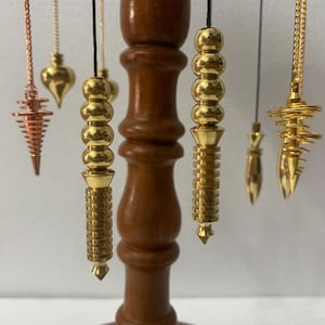 Peut inclure: Plusieurs pendules en laiton de formes variées suspendues à un support en bois. Les pendules présentent des formes coniques, en goutte d'eau et cylindriques. Le support est de couleur marron foncé, sur fond blanc uni.