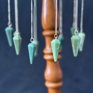 May include: A collection of green gemstone pendulums hanging from silver chains. The pendulums are faceted and cone-shaped, with a polished finish. They are displayed on a wooden stand against a dark blue background.