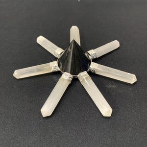 Puede incluir: Un generador de energía en forma de estrella con un centro negro en forma de cono y ocho varitas de cristal de cuarzo transparente. Las varitas están unidas al centro con detalles metálicos plateados. El objeto está sobre un fondo oscuro.