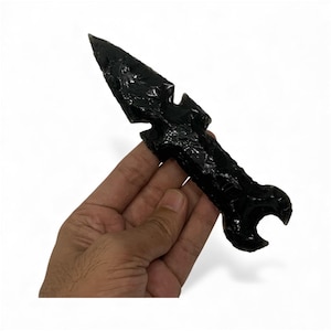 May include: A hand-carved, obsidian arrowhead with a sharp point and a textured surface. The arrowhead is a deep, glossy black and features a unique, symmetrical design. The arrowhead is held against a white background.