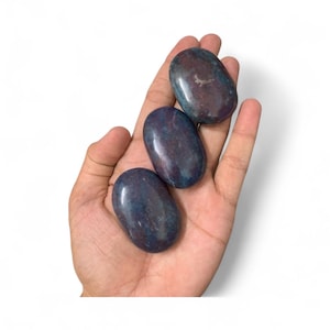 Op de afbeelding: Drie gepolijste, ovaalvormige stenen in blauwe en bruine tinten worden in een open hand gehouden. De stenen hebben een glad, glanzend oppervlak en variëren enigszins in kleur. De achtergrond is effen wit.