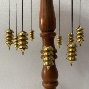 Puede incluir: Un carillón de viento de madera con múltiples niveles de campanas de metal dorado suspendidas de cuerdas negras. El poste de madera es de color marrón oscuro. Las campanas tienen un diseño en capas y redondeado con una parte inferior puntiaguda.