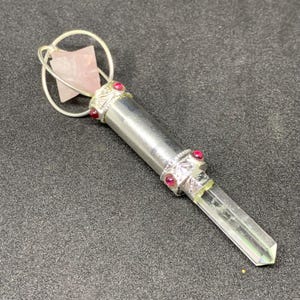 May include: A silver-colored crystal wand with a clear quartz point and a pink rose quartz at the top. The wand is adorned with red gemstones and decorative silver bands. The wand is designed with a loop at the top.