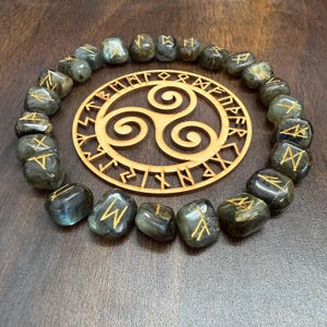 May include: A collection of labradorite rune stones with gold-coloured symbols arranged around a wooden Celtic Triskele symbol. The stones are a mix of grey and green, and the wooden symbol is light brown.