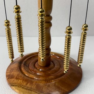 Op de afbeelding: Een houten pendelstandaard met een ronde basis en een centrale kolom. Vijf goudkleurige pendels hangen aan zwarte touwtjes. De pendels hebben een cilindrisch ontwerp met een reeks ribbels en decoratieve finials.