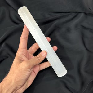 May include: A clear, white selenite wand, cylindrical in shape, held in a hand. The wand has a smooth, polished appearance and is approximately 20 cm long. The background is a dark, textured fabric.