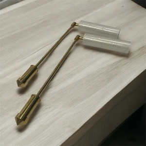 May include: Two brass plumb bobs with long, slender rods and pointed tips. The bobs have a cylindrical body and are resting on a light-colored wooden surface. Two wrapped cylindrical objects are also present.