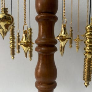 May include: A collection of gold-colored pendulums hanging from gold chains, with a dark brown wooden stand in the background. The pendulums have various shapes, including teardrop and conical designs. The background is a plain white wall.