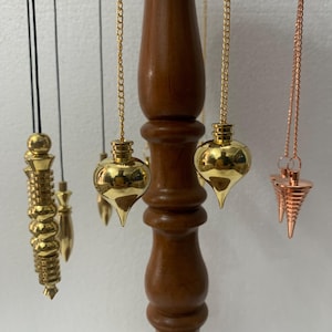Könnte beinhalten: Eine Sammlung von Pendeln in verschiedenen Formen und metallischen Ausführungen, darunter Gold und Kupfer, die an einem Holzständer hängen. Die Pendel sind an Ketten und Kordeln befestigt und zeigen ihre unterschiedlichen Designs.