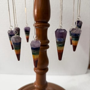 Op de afbeelding: Een verzameling van zeven chakra kristallen pendels die aan een houten standaard hangen. Elke pendel heeft een facetvormige, langwerpige vorm met kleurlagen die de chakra's vertegenwoordigen: rood, oranje, geel, groen, blauw, indigo en violet. De pendels hangen aan zilveren kettingen.