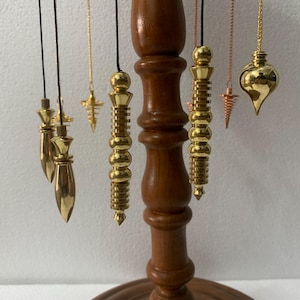 Puede incluir: Un soporte de madera exhibe varios péndulos dorados de diversas formas, suspendidos de cuerdas negras. Los péndulos incluyen diseños de lágrima, cilíndricos y de cruz. El soporte es de color marrón oscuro, sobre un fondo blanco liso.