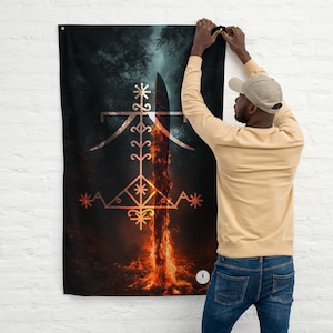 May include: A black wall flag with a stylized design featuring a central flame and intricate symbols. The design is predominantly white and gold against a dark background, with a person hanging the flag.