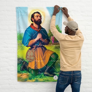 May include: A colorful religious flag featuring a kneeling figure of a saint with a beard, wearing a blue robe and a yellow skirt. The saint is holding his hands in prayer. The flag is hanging on a white wall.