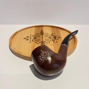 Könnte beinhalten: Eine braune Tabakpfeife mit gebogenem Stiel und goldenem Emblem. Die Pfeife liegt auf einem runden Holztablett mit einem geätzten Design. Die Pfeife hat eine dunkelbraune Oberfläche und ein schwarzes Mundstück.