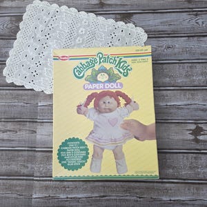 May include: A vintage Cabbage Patch Kids Paper Doll set. The box features a doll illustration with red pigtails and a pink dress. The box also includes the text "PAPER DOLL" and "AGES 4 THRU 8".