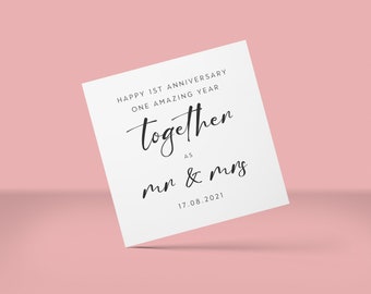 May include: A white card with the text "Happy 1st Anniversary, One Amazing Year, together as mr & mrs, 17.08.2021" in black script. The card is angled against a soft pink background, creating a shadow. The card is a simple, elegant design.