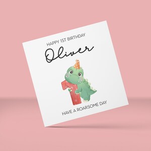May include: A white birthday card with the text "HAPPY 1ST BIRTHDAY Oliver" in black script. A green dinosaur wearing a party hat and holding a red number one is featured. The card also says "HAVE A ROARSOME DAY".