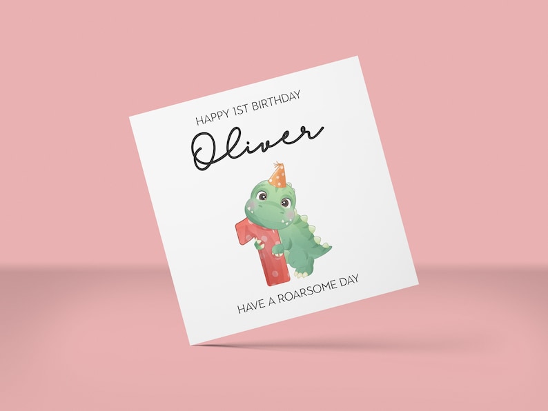 May include: A white birthday card with the text "HAPPY 1ST BIRTHDAY Oliver" in black script. A green dinosaur wearing a party hat and holding a red number one is featured. The card also says "HAVE A ROARSOME DAY".
