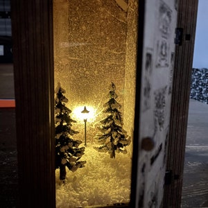 Op de afbeelding: Een houten lantaarn met een besneeuwde wintersc&egrave;ne erin. De sc&egrave;ne toont twee kleine dennenbomen en een lantaarnpaal, die allemaal worden verlicht door een warm geel licht.