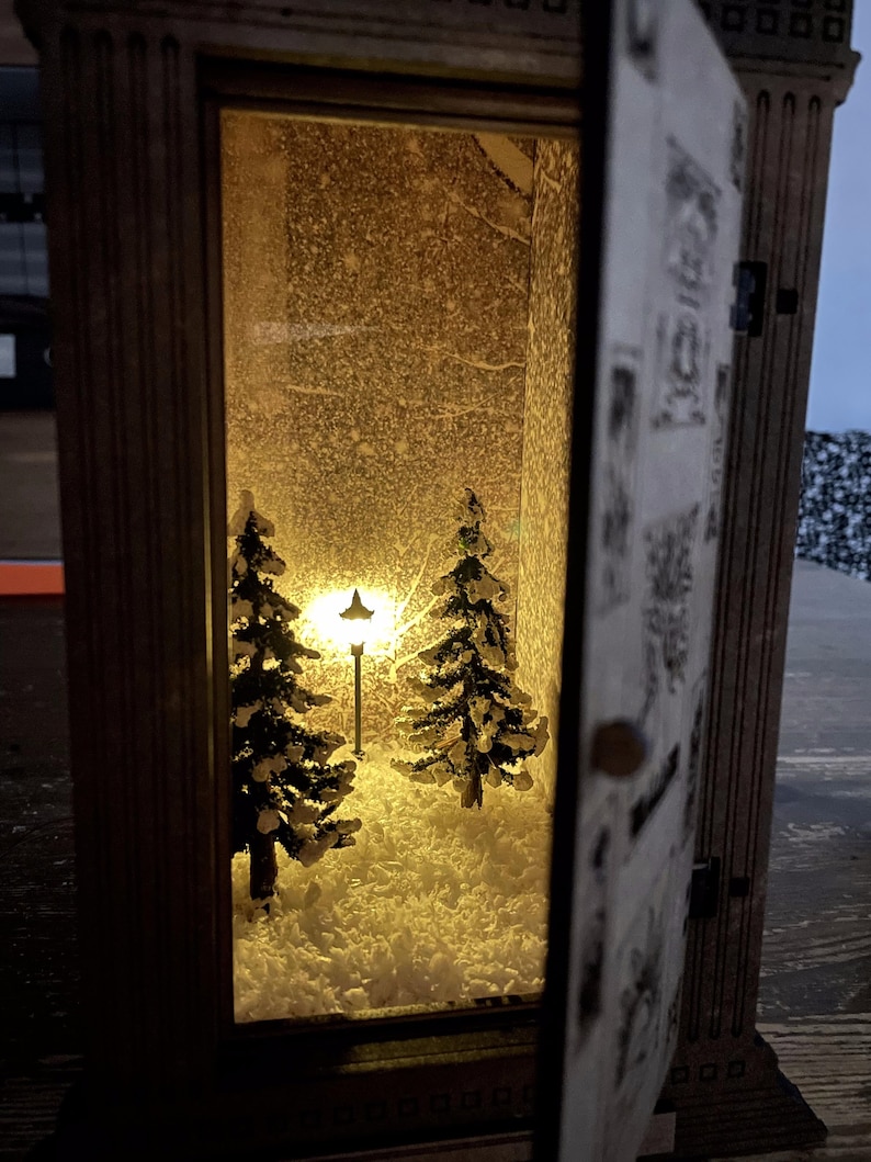 Op de afbeelding: Een houten lantaarn met een besneeuwde wintersc&egrave;ne erin. De sc&egrave;ne toont twee kleine dennenbomen en een lantaarnpaal, die allemaal worden verlicht door een warm geel licht.