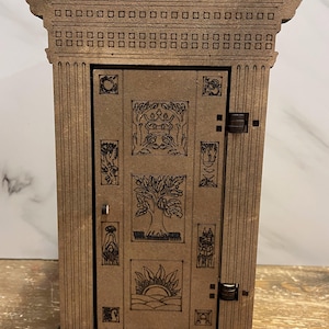 Op de afbeelding: Een houten doos met een decoratieve deur met ingewikkelde houtsnijwerk van bomen, kronen en andere symbolen. De doos heeft een bruine afwerking en een eenvoudig ontwerp.