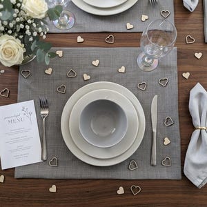 May include: A dining table set for a special occasion, featuring grey placemats, white plates, and grey bowls. Silver cutlery and wine glasses are placed around the plates. Wooden heart confetti and a menu card are scattered. A bouquet of white roses is in the background.