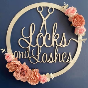 Puede incluir: Un círculo de madera blanco con las palabras "Locks and Lashes" en escritura cursiva. Un par de tijeras plateadas está por encima del texto. El círculo está decorado con flores rosas y blancas.