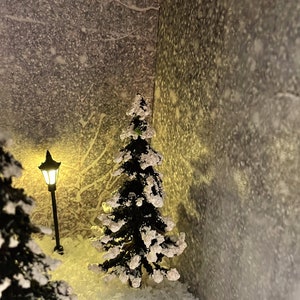 Op de afbeelding: Een besneeuwde wintersc&egrave;ne met twee kleine, met sneeuw bedekte dennenbomen en een verlichte lantaarnpaal. De achtergrond is een getextureerde, grijze muur.